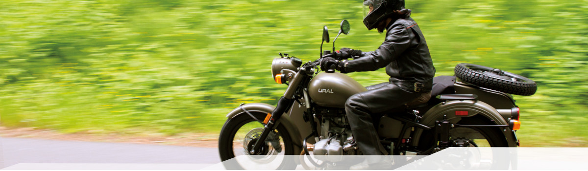 Helmeted person driving a dark tan Ural motorcycle quickly down a road lined by trees.